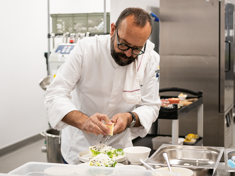 A man wearing a white chef's coat stands grating cheese over a dish in a kitchen setting.