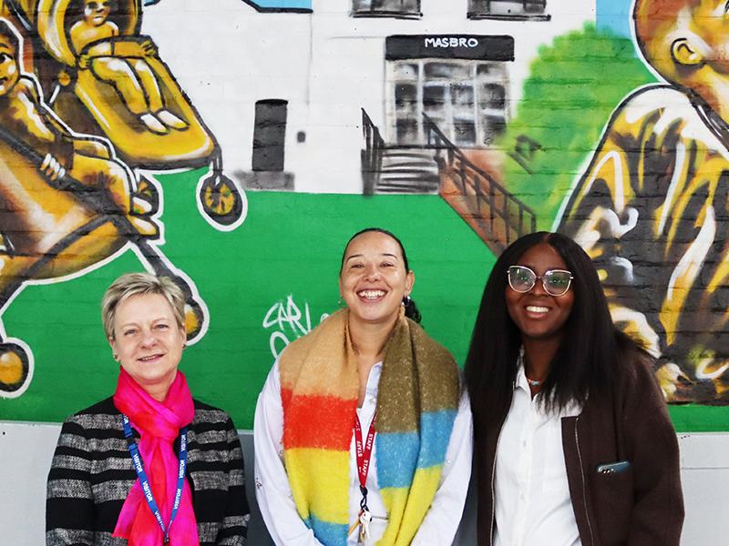 In front of a striking mural, three women stand together.