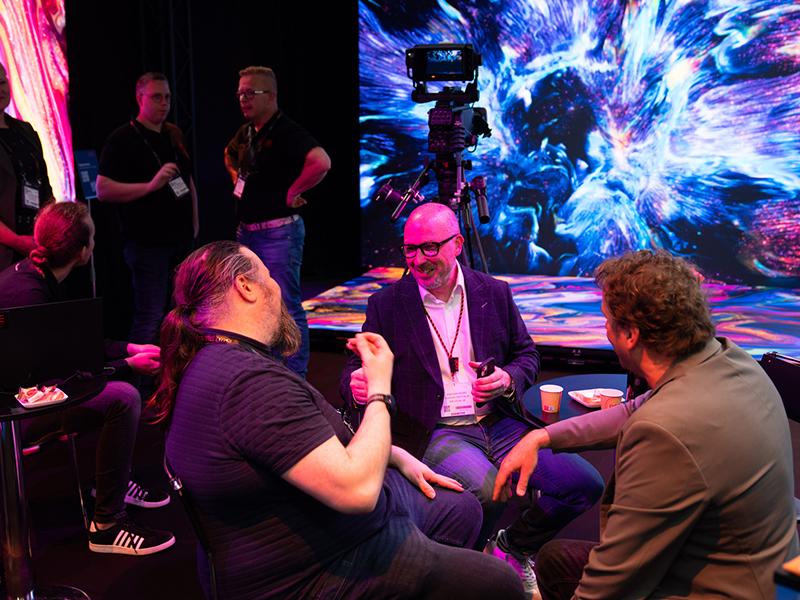 A group of people engage in conversation and networking in front of a colourful, swirling digital backdrop and high-tech camera equipment at a trade show or exhibition.