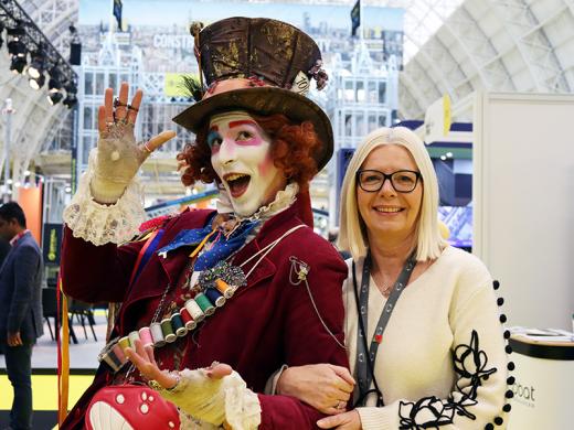 A woman with glasses stands beside a man dressed as mad hatter, both smiling in a lively setting.