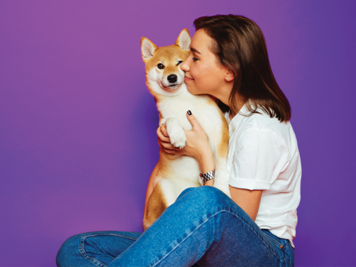 A young woman with long dark brown hair is sitting on the floor, wearing a white t-shirt and jeans, and cuddling a Shiba Inu dog.