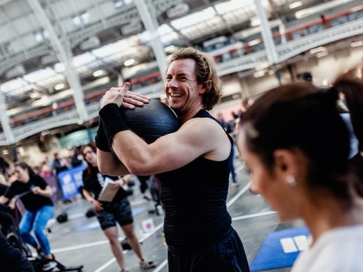 A man with shoulder-length hair, wearing a black tank top and arm sleeves, smiles while holding a heavy black medicine ball close to his chest.