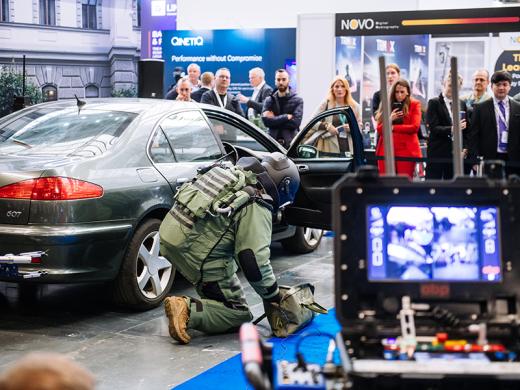 A person in military gear kneels beside a green car during a demonstration at a trade show.