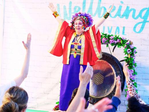  A woman dressed in a vibrant red and purple outfit with a decorative headdress leads an interactive session at the Wellbeing Festival, raising her hands as the audience follows along.