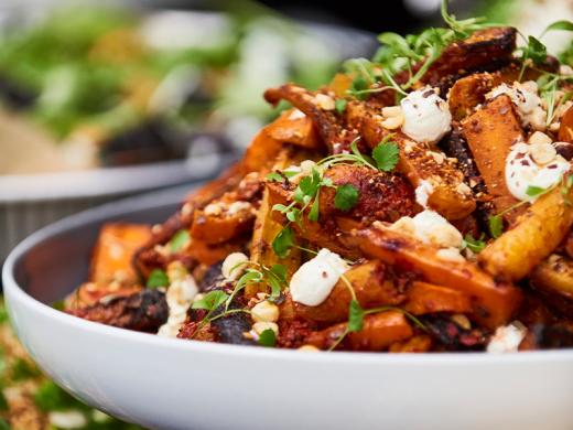 A close-up of a vibrant dish served in a white bowl, featuring roasted vegetables, possibly sweet potatoes or carrots, garnished with crumbled cheese, fresh herbs, and chopped nuts. 