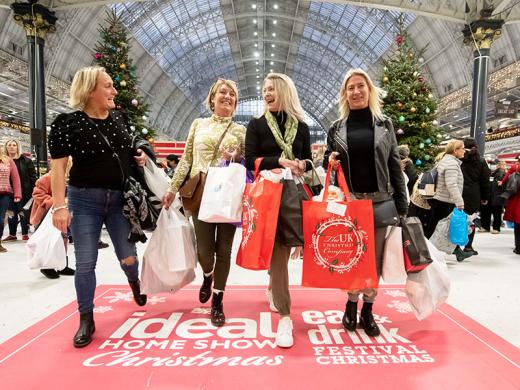 Four individuals carrying shopping bags walk along the red carpet at the Ideal Home Show at Christmas event in a large, decorated hall.