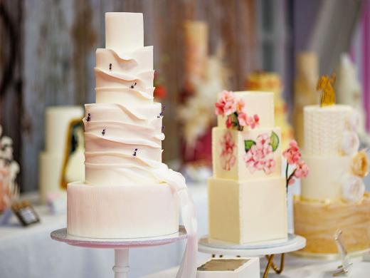 Assorted elegant cakes, including a tall, ribbon-wrapped cake and others adorned with flowers and bees, displayed at a bakery event.