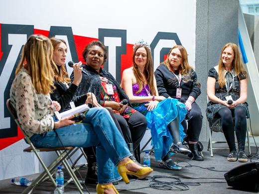 Five panelists engaged in a discussion, seated in front of a large sign.