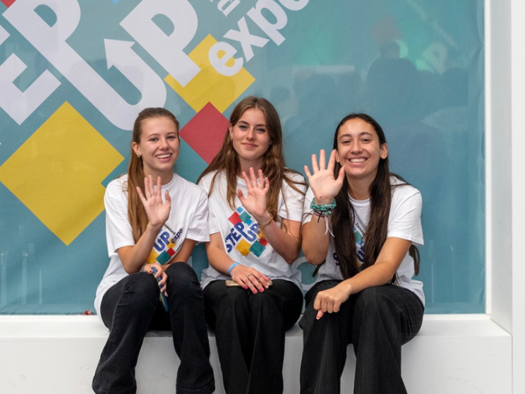 Three girls waving to camera and sitting closely together on a white bench.