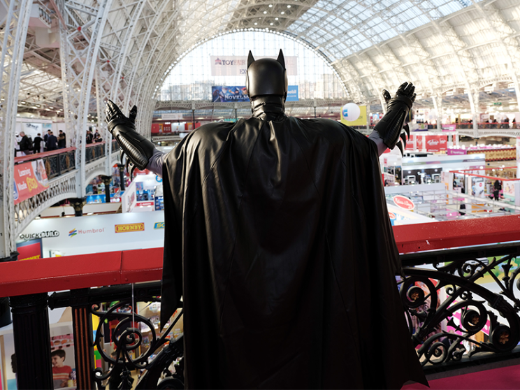 An individual dressed as Batman stands heroically, showcasing his iconic cape and cowl while overlooking an exhibition show floor.