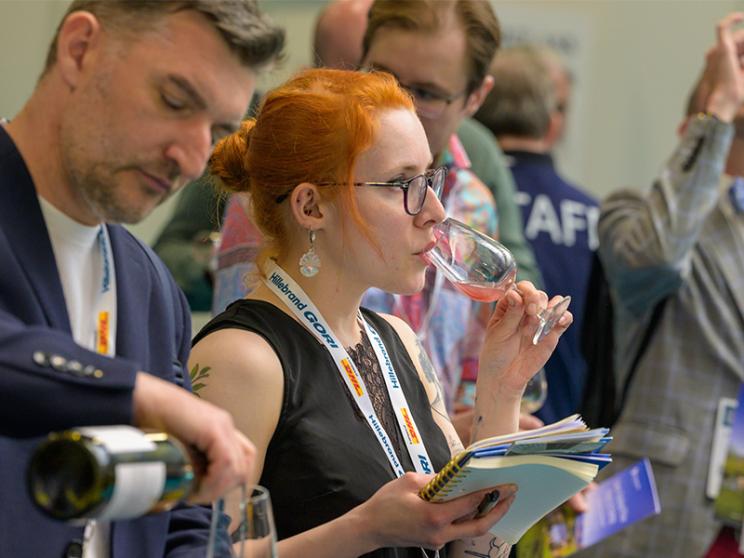 A woman sipping on a glass of wine while a man standing next to her pours wine into a glass.