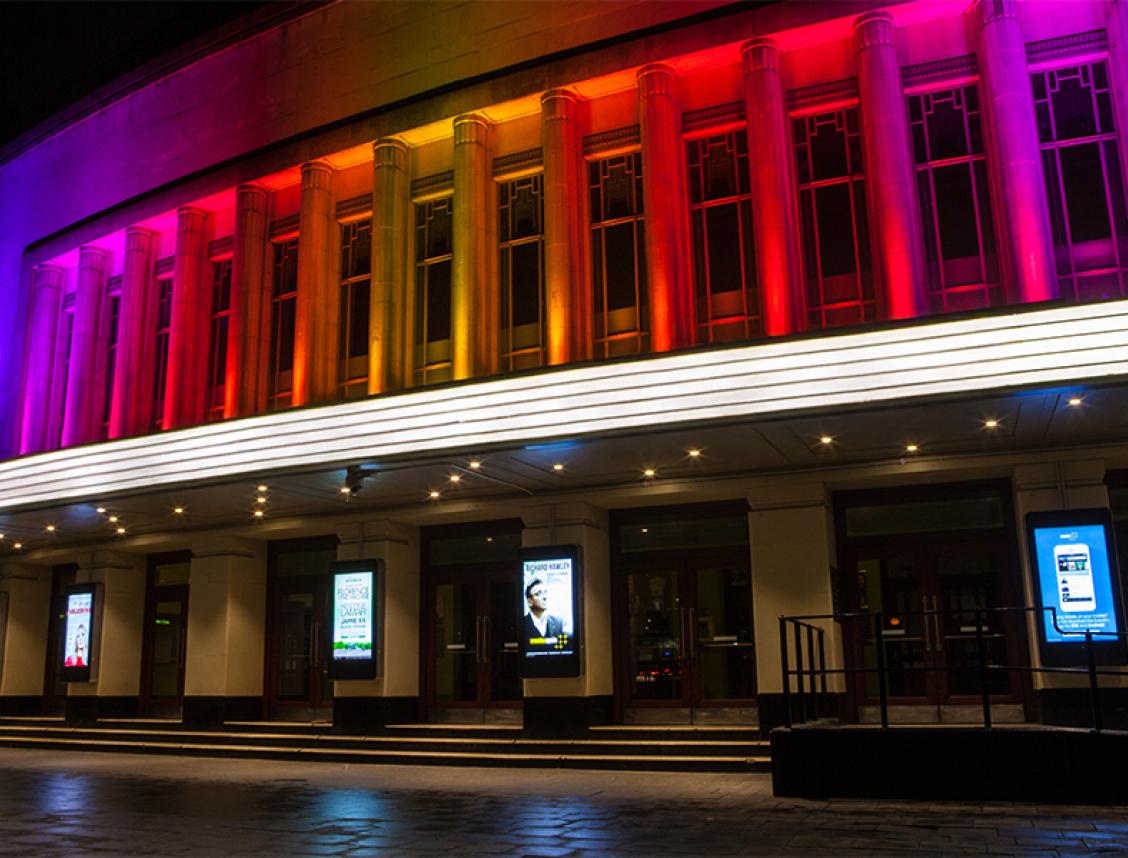 The front of a historic London venue lit in rainbow colours at night.
