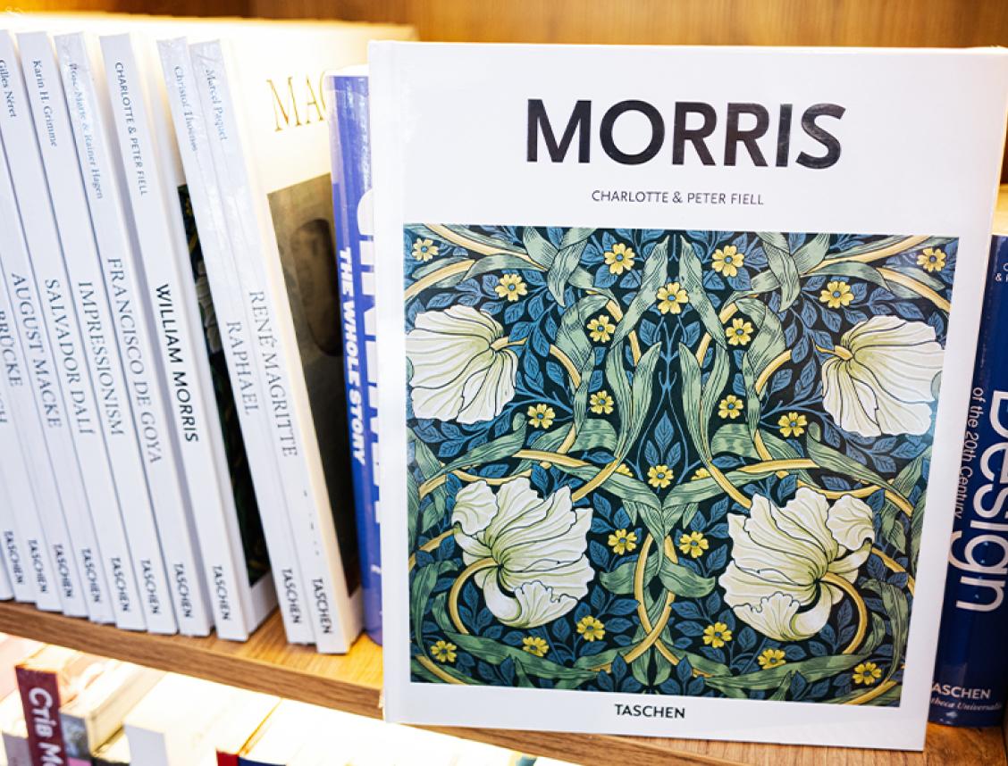A wooden bookshelf filled with art and design books, prominently featuring a “William Morris” title with a floral patterned cover by Charlotte and Peter Fiell.