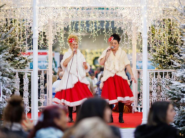 Two performers in traditional red and white outfits and fur shawls are dancing and laughing on a stage decorated with white lights and surrounded by Christmas trees.