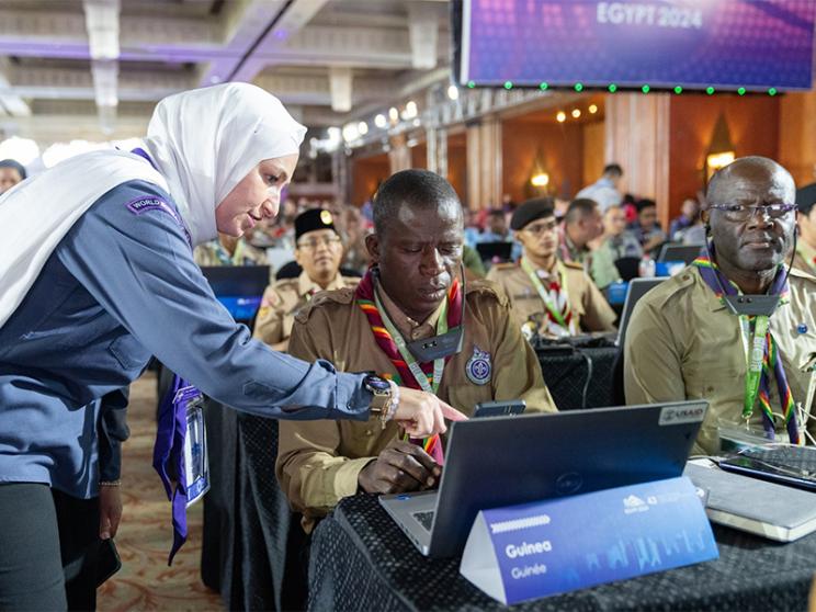 A woman in a Scouts uniform leans over to assist a delegate using a laptop at an international Scouting conference, with other attendees seated and working in the background.