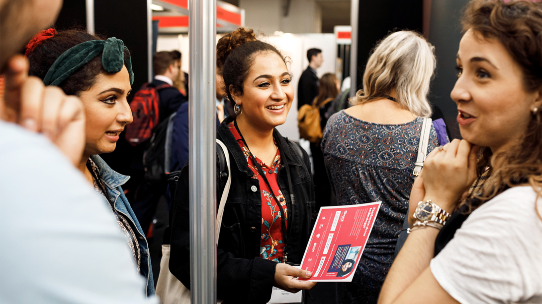 A diverse group of individuals gathered around a trade show booth, engaged in conversation at a lively event.