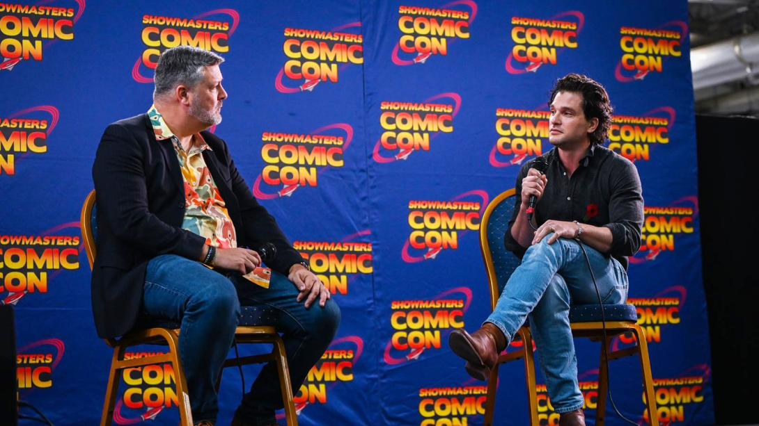 Two men seated on chairs at a comic con, engaged in conversation.