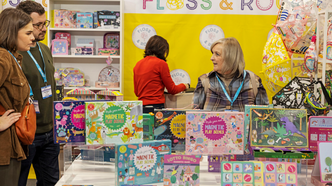 A woman and a couple looking at board games set up on a table at a busy tradeshow.