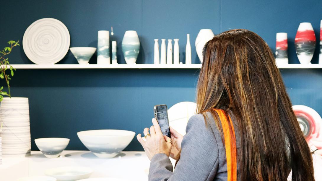 A woman stands in front of an art showcase whilst taking a picture on her phone.