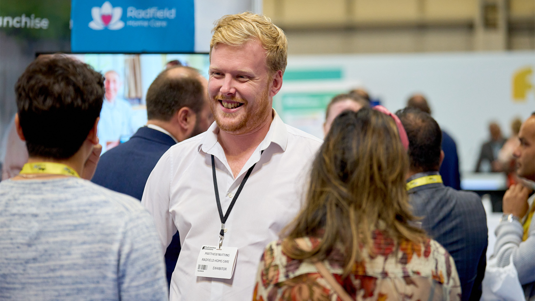  A smiling man at a trade show, surrounded by engaged attendees.