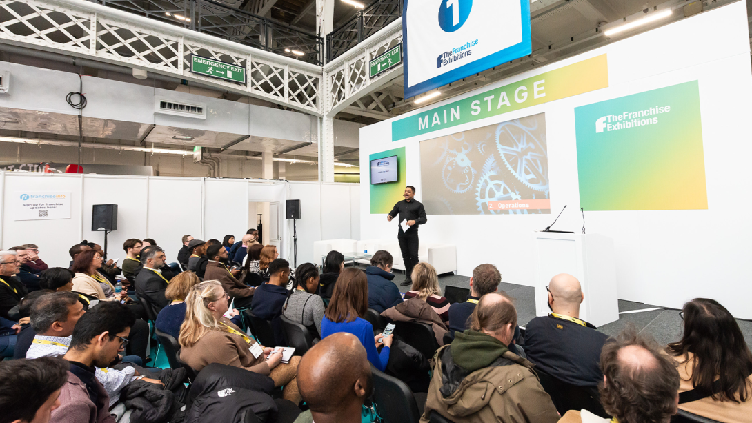 A presenter speaking on stage, speaking to a large audience at a trade show.