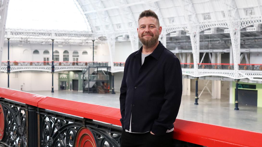 A man stands on a balcony in an indoor space, gazing thoughtfully into the distance.