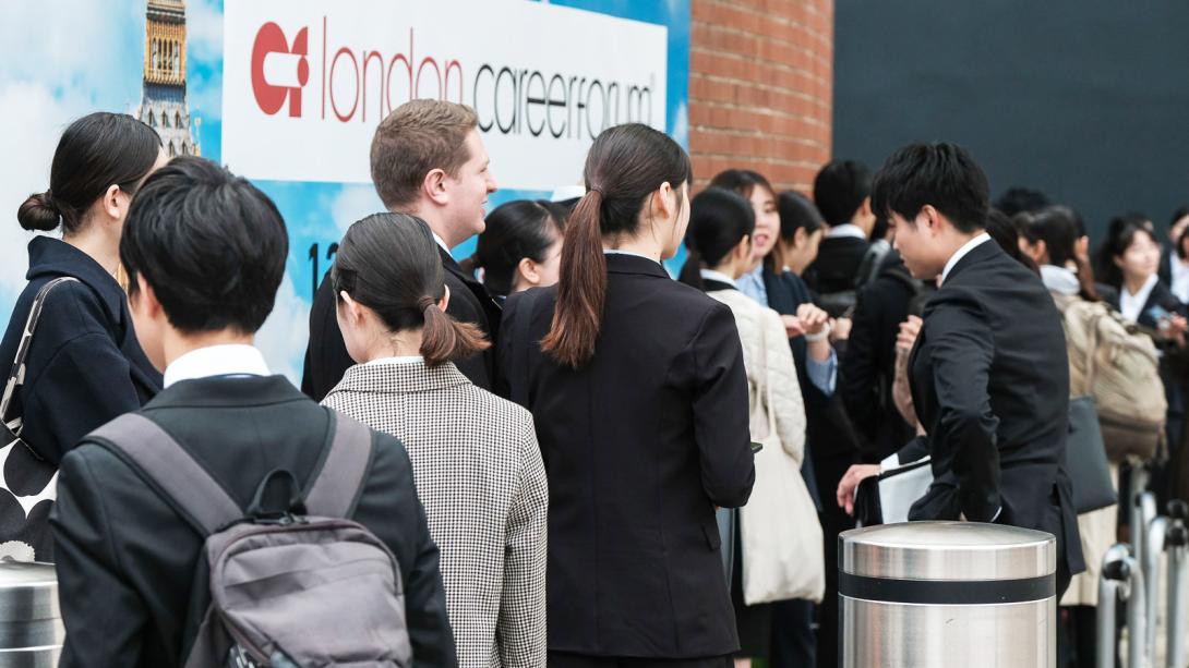 A diverse group of individuals standing in line, waiting patiently in an outdoor setting.