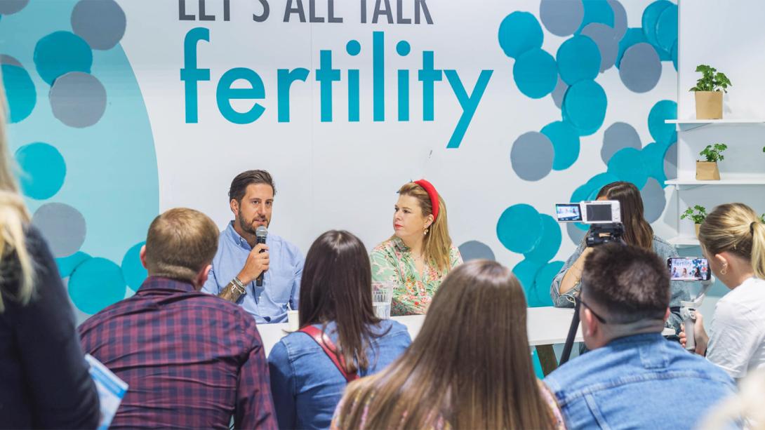 A diverse group of individuals seated in front of a sign reading "Let's All Talk Fertility," promoting open discussions on fertility.