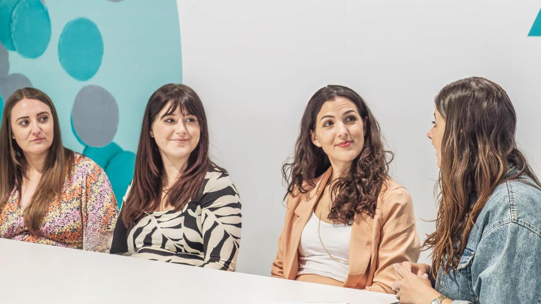 Four women engaged in conversation while seated around a table, sharing ideas and laughter in a lively discussion.