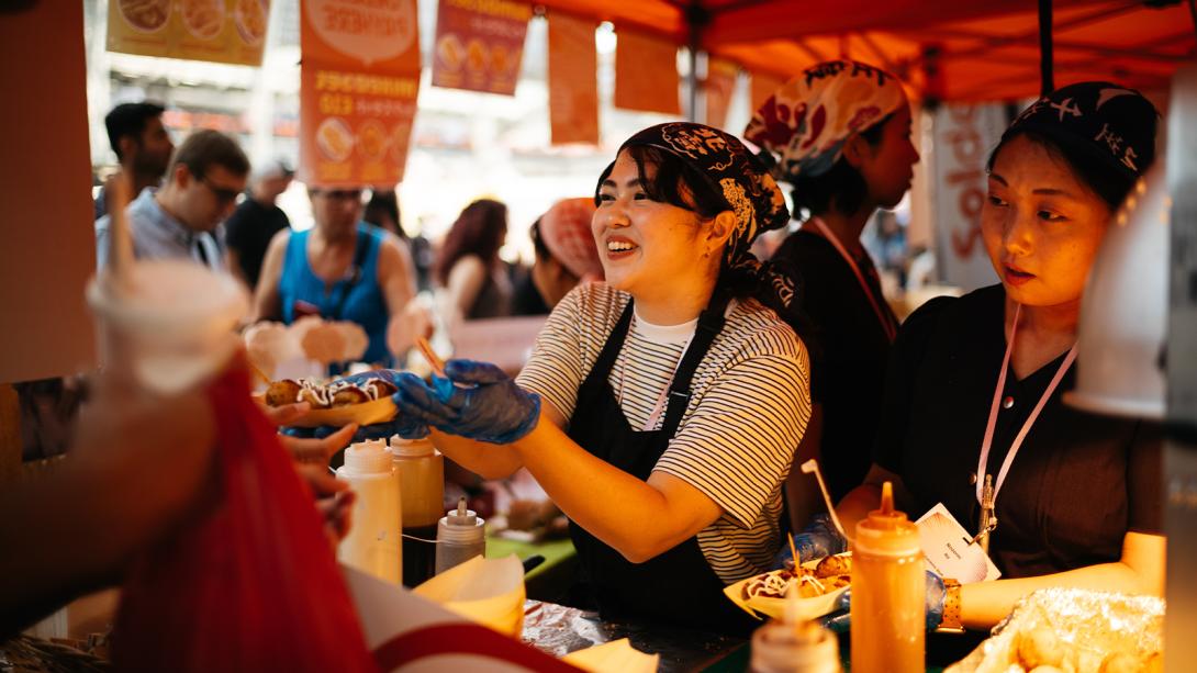  Two women serve food at a bustling food stand.