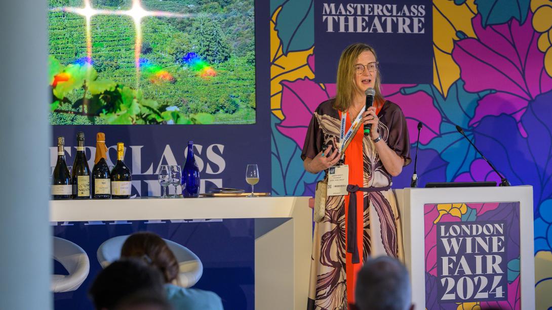 A woman stands at a podium, addressing attendees at a wine tasting event.