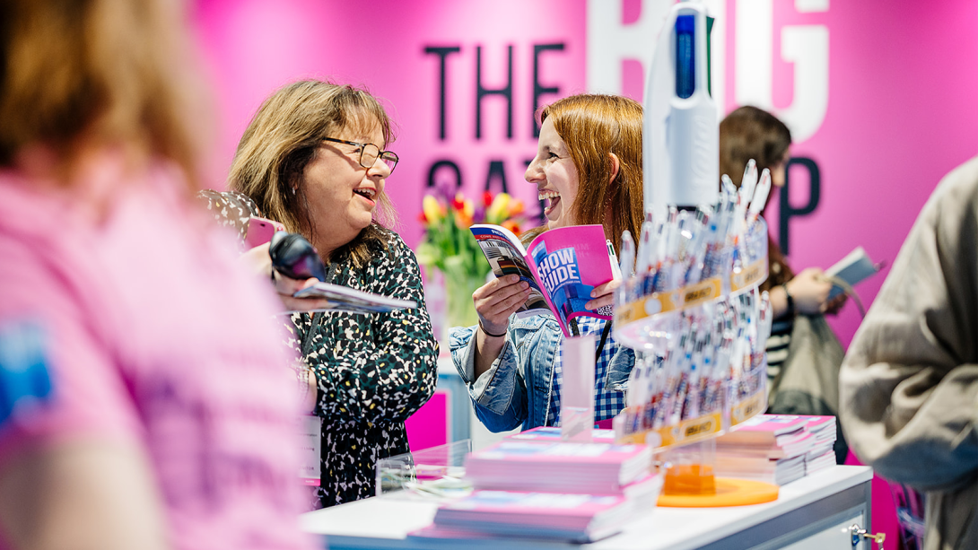 Two women smiling and laughing together at a vibrant expo booth.