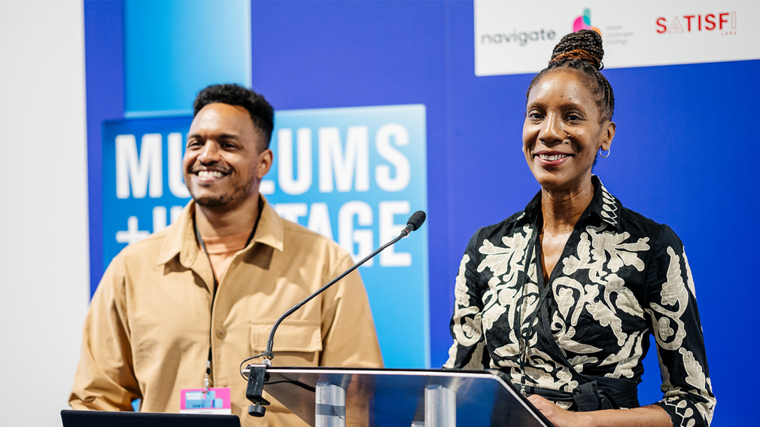 Two individuals at a podium, with a sign in the background that reads "Museums and Heritage".