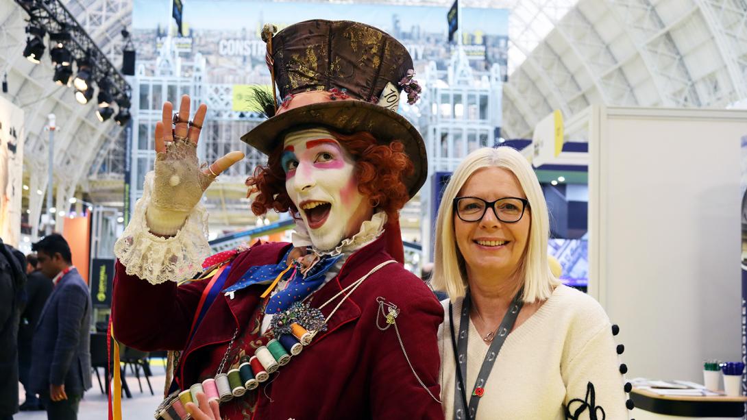 A woman with glasses stands beside a man dressed as a mad hatter, both smiling in a lively setting.
