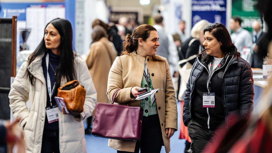  Women at a trade show, each holding various bags and purses, engaged in conversation.