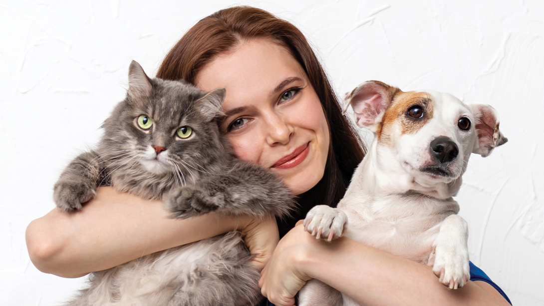 A young woman with brown hair smiles while holding a fluffy grey cat and a white and brown dog. 