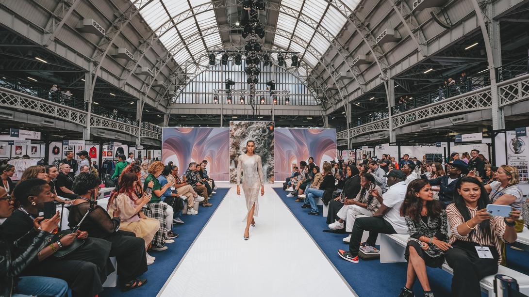 A woman confidently walks down a runway inside a spacious, modern building, showcasing her stylish outfit.