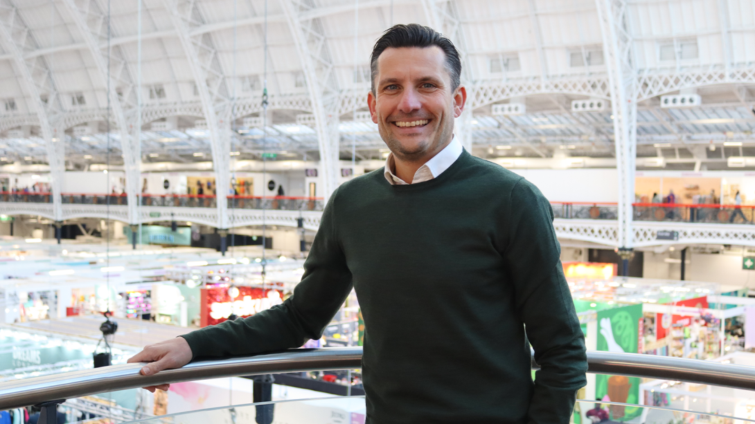 A smiling man wearing a green sweater and white shirt stands leaning on a railing in an indoor venue with a high arched ceiling and visible booths and displays in the background.