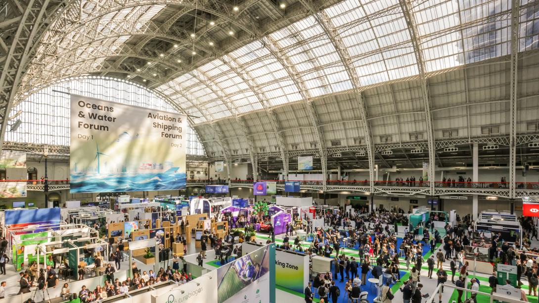 Panoramic view inside a large exhibition hall at, featuring numerous booths and attendees under a vast glass ceiling.