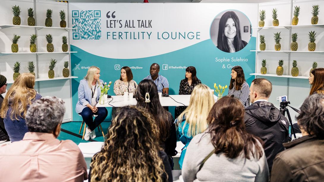 A diverse group of individuals seated at a table, with a wall sign reading "Fertility Lounge" in the background.