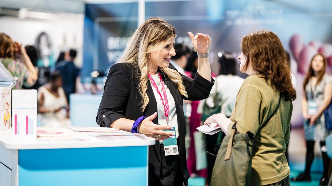 A woman engages in conversation with another woman at a bustling trade show, surrounded by various displays and attendees.