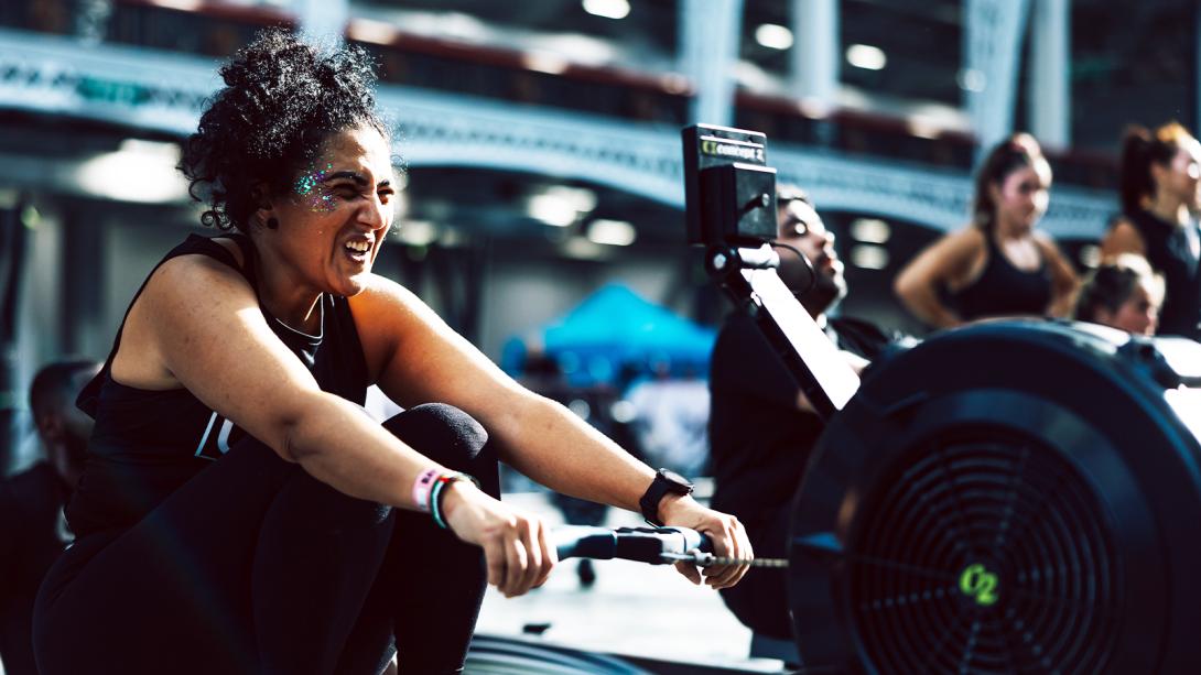 A woman with curly hair and glitter on her face is mid-rowing on an exercise machine.