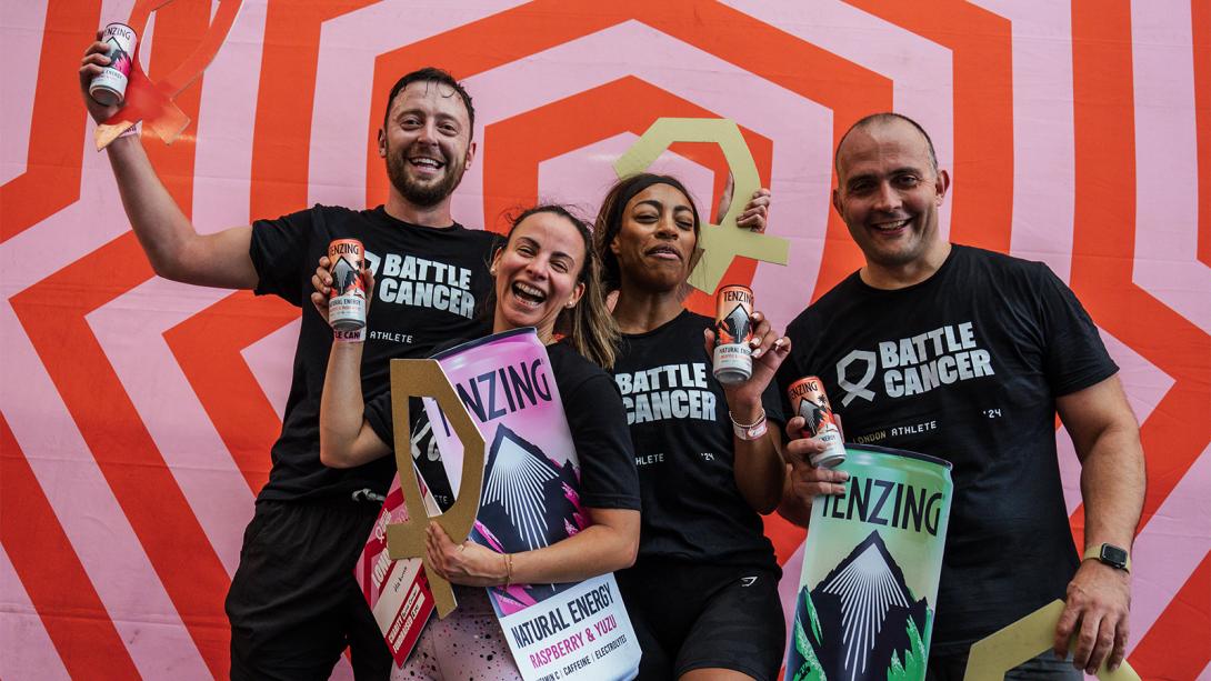 A group of four people wearing black "Battle Cancer" t-shirts celebrate in front of a bold red and pink geometric backdrop.