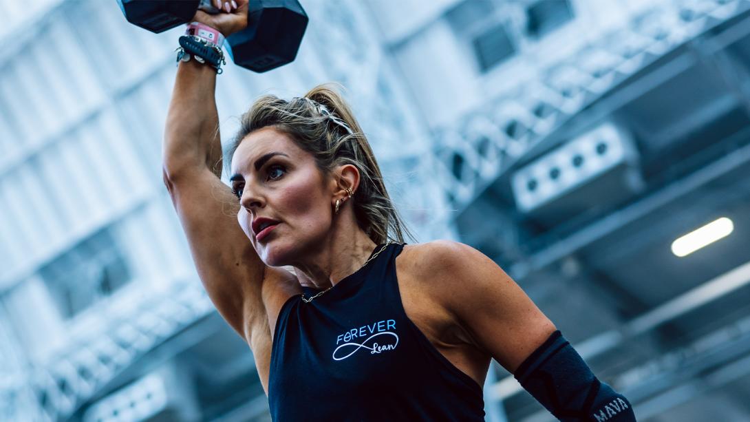 A determined-looking woman with a strong physique lifts a dumbbell overhead while competing in a fitness event.