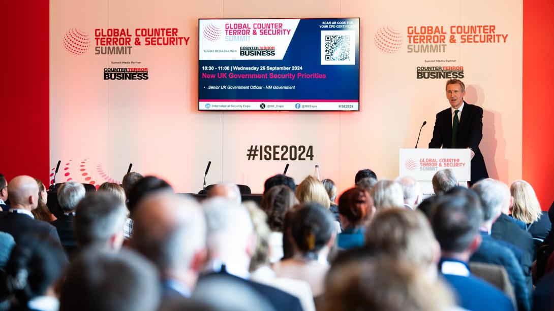 A person is speaking at the Global Counter Terror & Security Summit, standing at a podium with banners displaying the event name. The audience in the foreground is listening attentively.