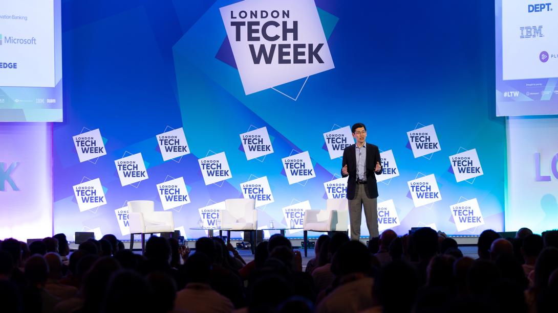 A person presenting on stage at London Tech Week, with logos of sponsors like Microsoft, Edge, IBM, and the Department of Trade visible in the background.