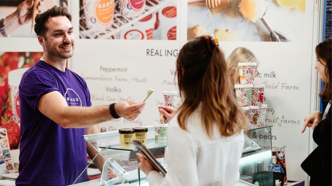 A man wearing a purple t-shirt stands behind a counter at a food stall, smiling as he hands a sample to a woman in a white coat.