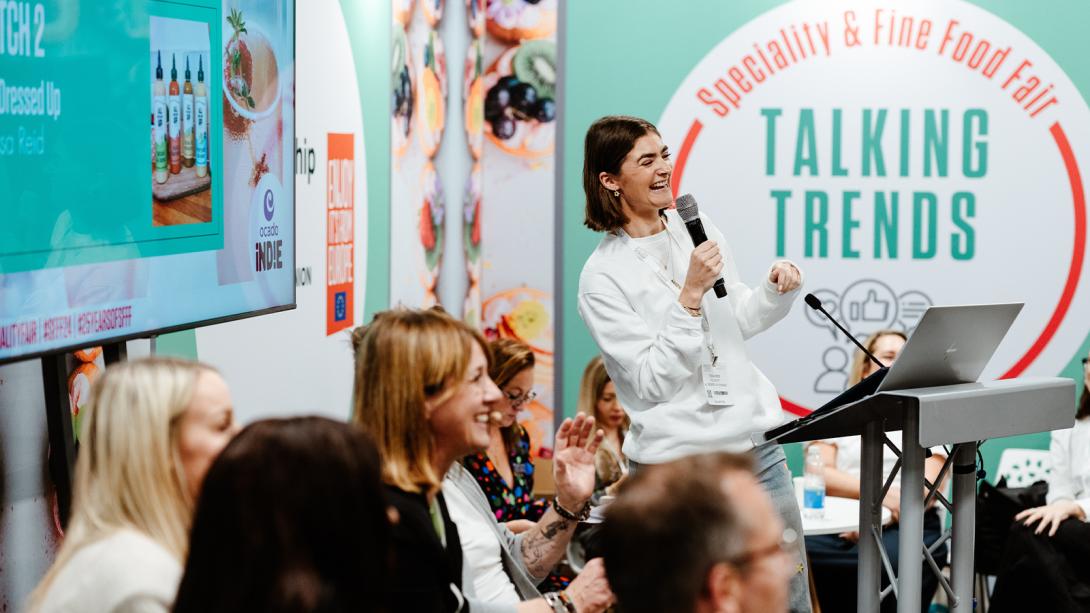 A woman in a white sweater stands at a podium, holding a microphone and laughing while speaking to an engaged audience.