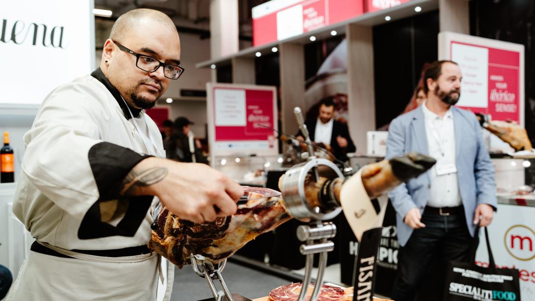 A chef wearing glasses, a white apron, and black-trimmed sleeves slices a large piece of cured meat at a speciality food exhibition. 