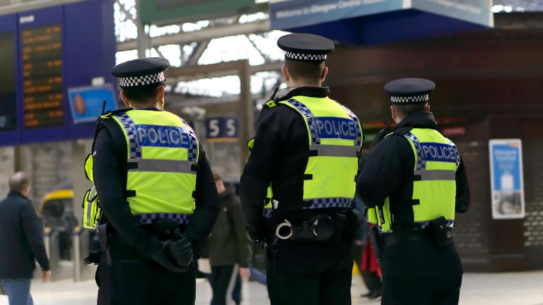 Three uniformed police officers wearing high-visibility vests stand inside a busy train station.